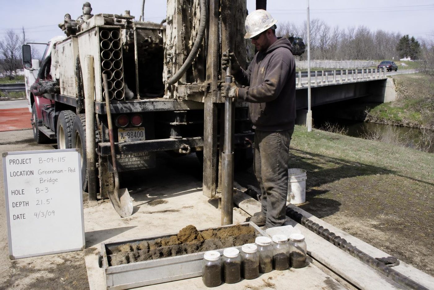 Truck-mounted drill rig taking a soil boring beside a bridge, with sample jars in the foreground.