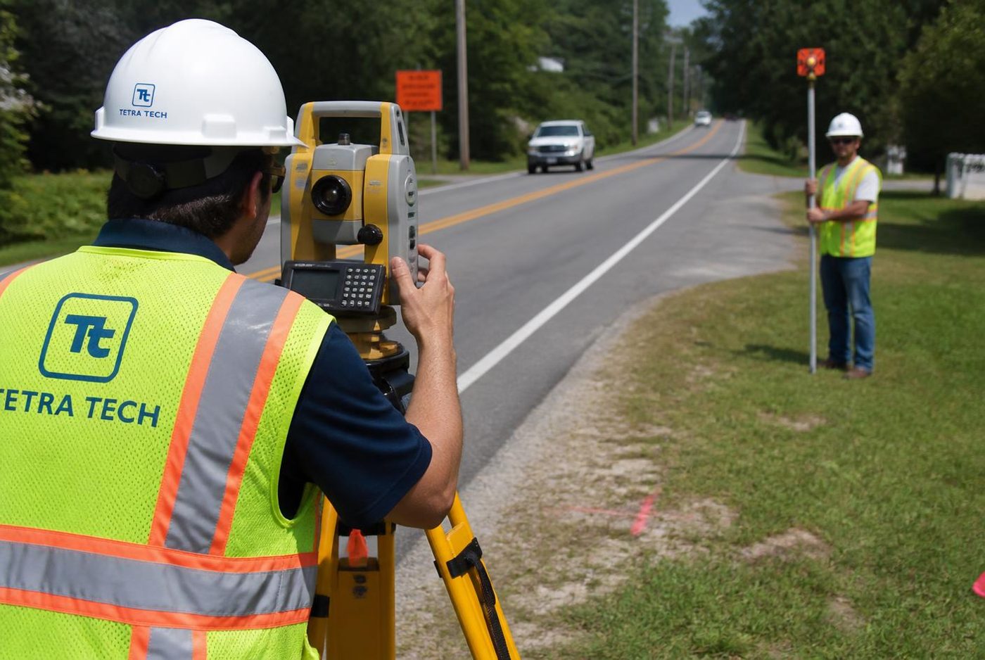 Surveyor operating a total station beside a rural highway with a rod person downstream.
